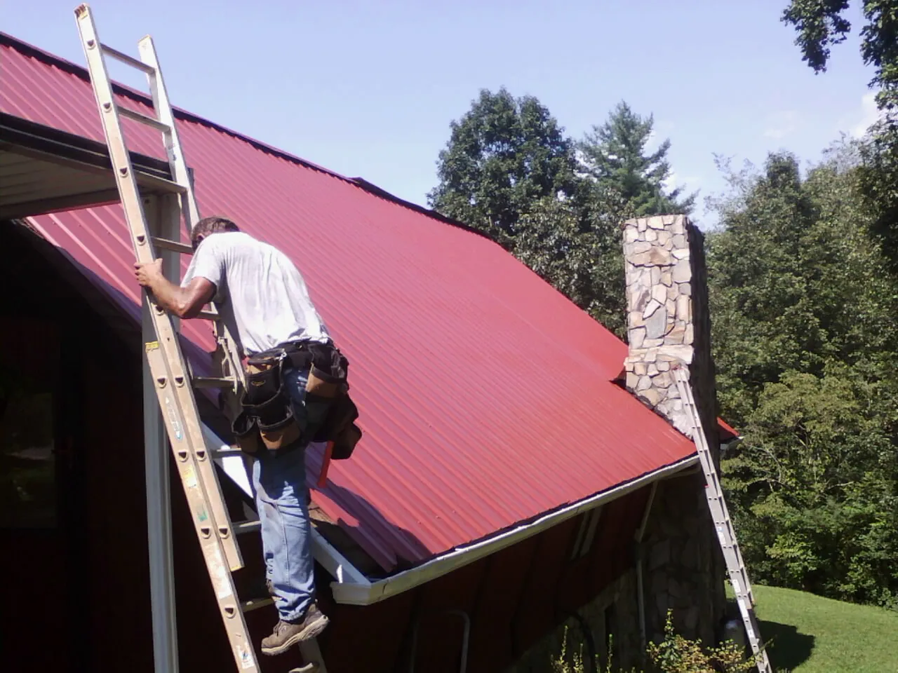 Roofer on a ladder beside a red metal roof and stone chimney — Soffit & Fascia Repair by Roofing Brothers in Idaho Springs